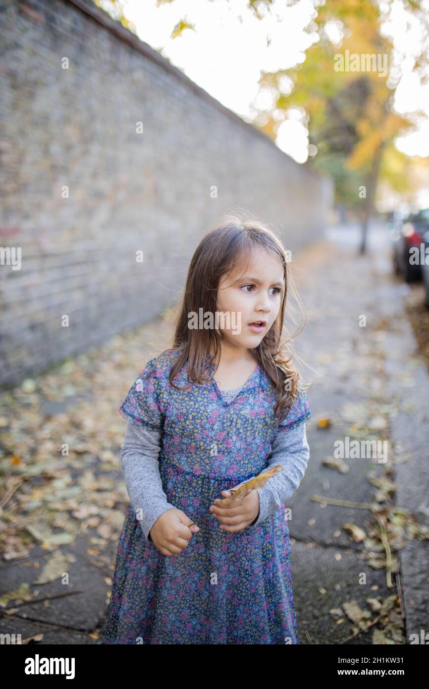 Ritratto stile immagine di una piccola bruna ragazza guardando la distanza e in piedi accanto ad un muro di mattoni e sul marciapiede coperto in autunno lasciare Foto Stock