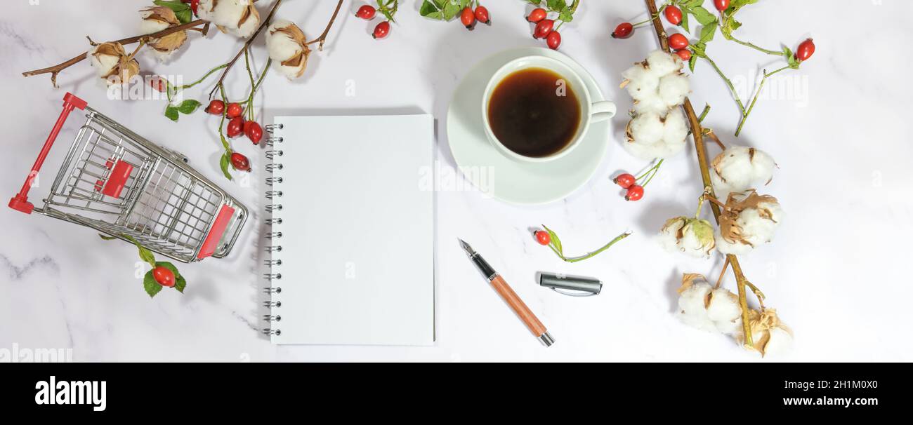 carrello, taccuino bianco e tazza di caffè su un tavolo di marmo chiaro con fianchi di rosa e rami di cotone, decorato per una lista in vendita autunnale, Foto Stock