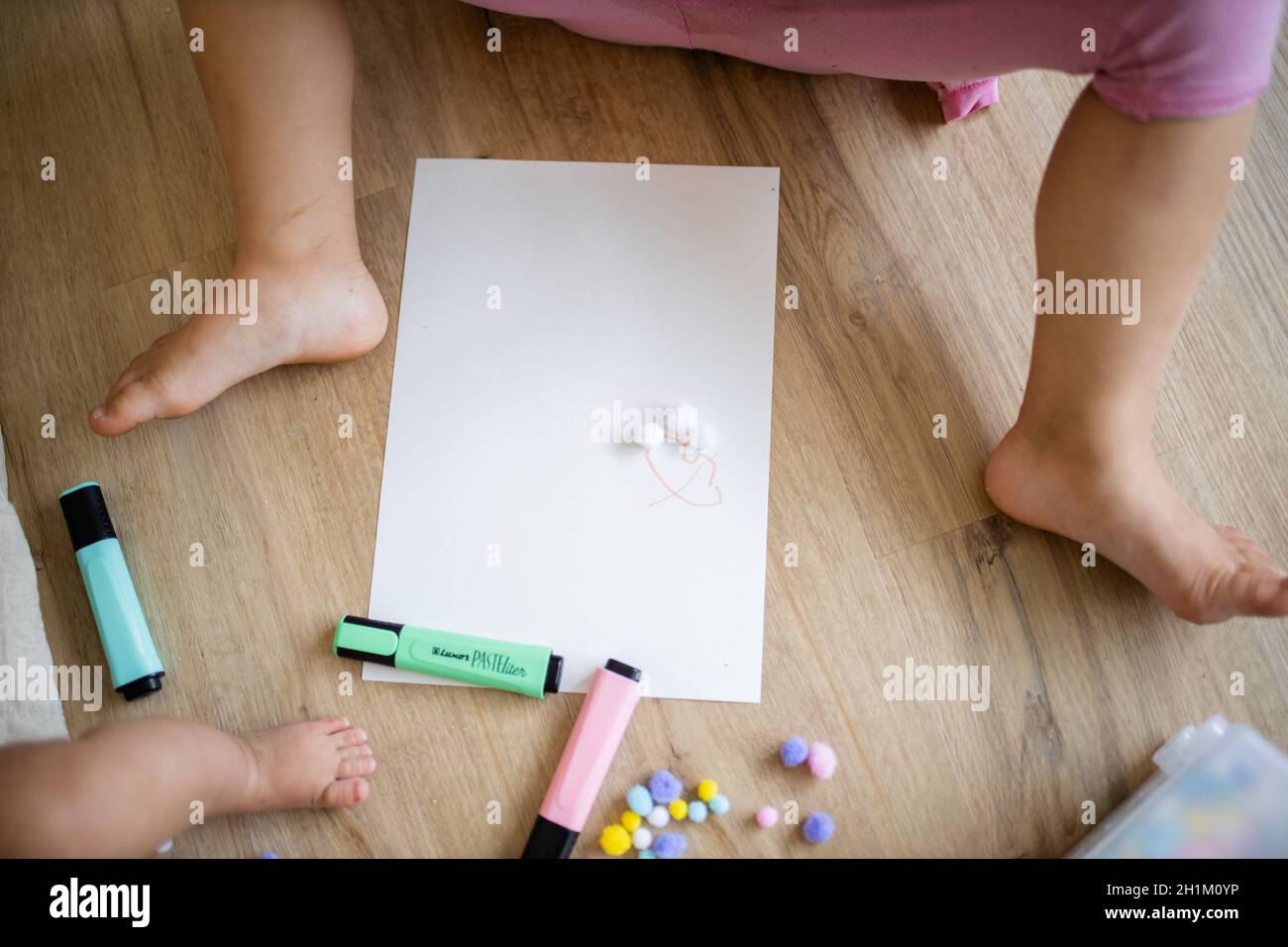 Immagine di un foglio di carta bianca, di un pennarello e di palline di cotone colorate su un pavimento di legno lungo la gamba di un bambino e le gambe di una littl Foto Stock