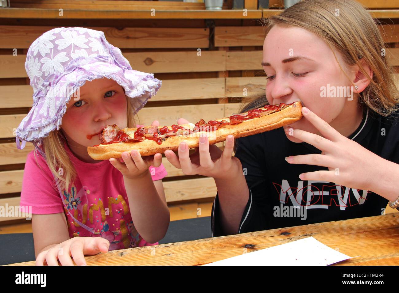 Due ragazze mangiano un panino grande allo stesso tempo. Due sorelle mangiano un delizioso panino su entrambi i lati. Cena fast food. I bambini dividono un panino. Foto Stock