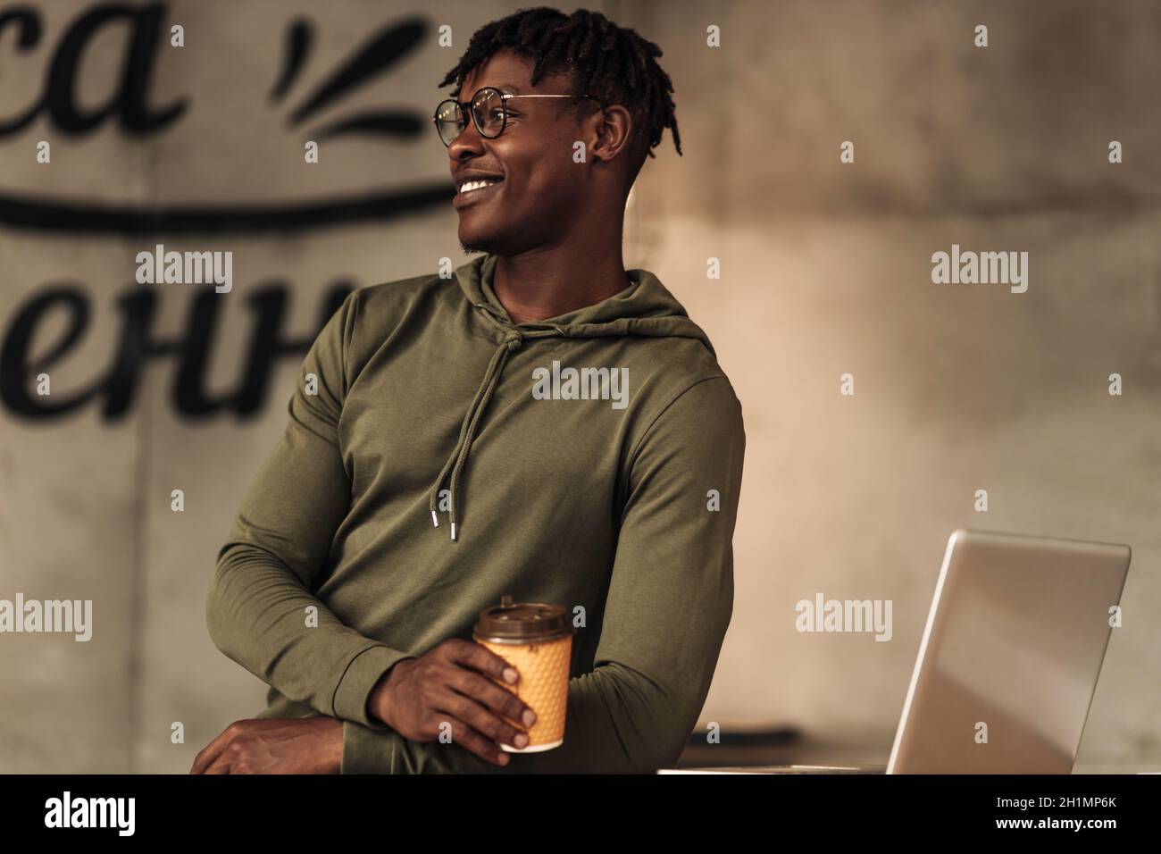 allegro uomo africano con una tazza di carta bianca nelle sue mani, seduto in un caffè, sorridente e guardando la macchina fotografica. dipendente al caffè o tè. aroma di co Foto Stock