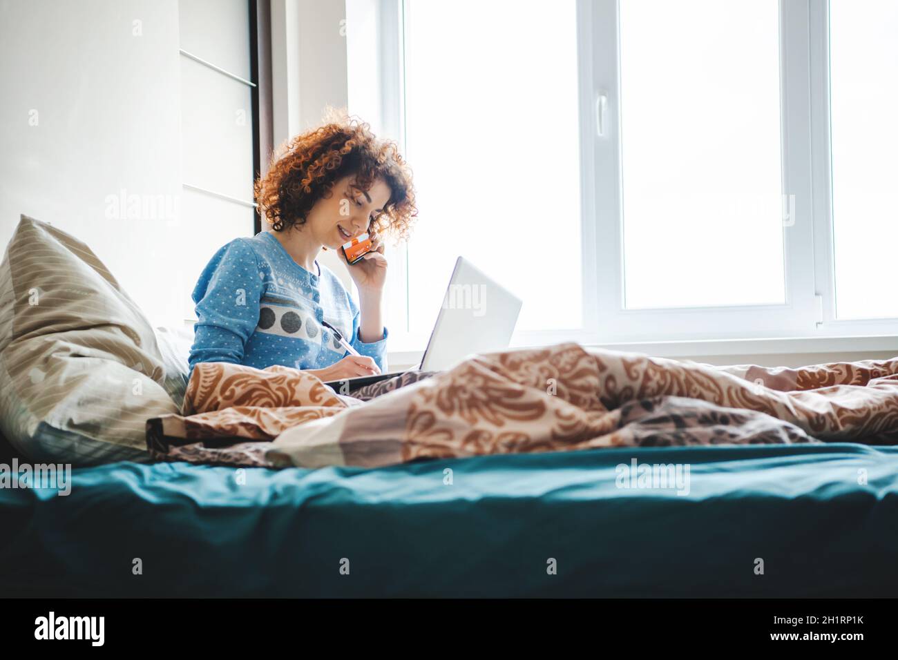 Donna curly che parla al telefono direttamente dal letto notando i compiti. Lezioni online Foto Stock