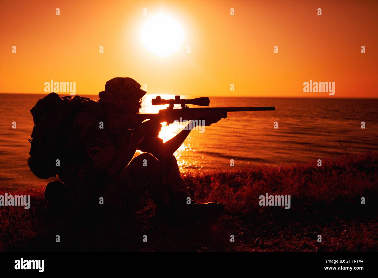 Il cecchino della squadra di Commando, lo sparatutto dell'esercito con forze speciali che punta, spara il fucile da cecchino mentre si siede sul mare o sulla riva dell'oceano durante il tramonto. Costa o confine gua Foto Stock