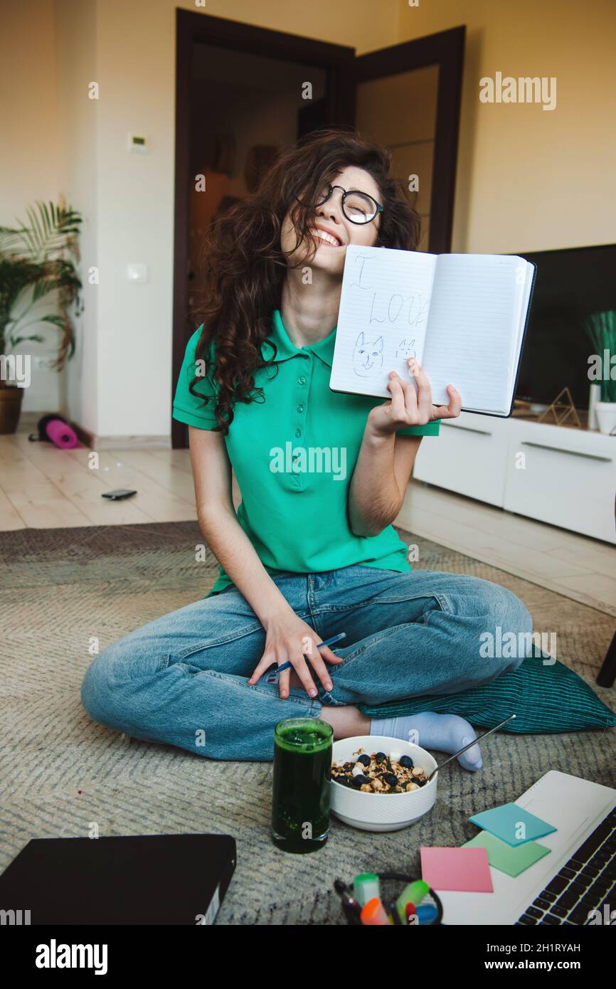 Donne che si divertono nella sua stanza seduto sul pavimento e tenendo un notebook aperto nella sua mano sinistra, puntando alla macchina fotografica che ama i gatti. Sorridente Foto Stock