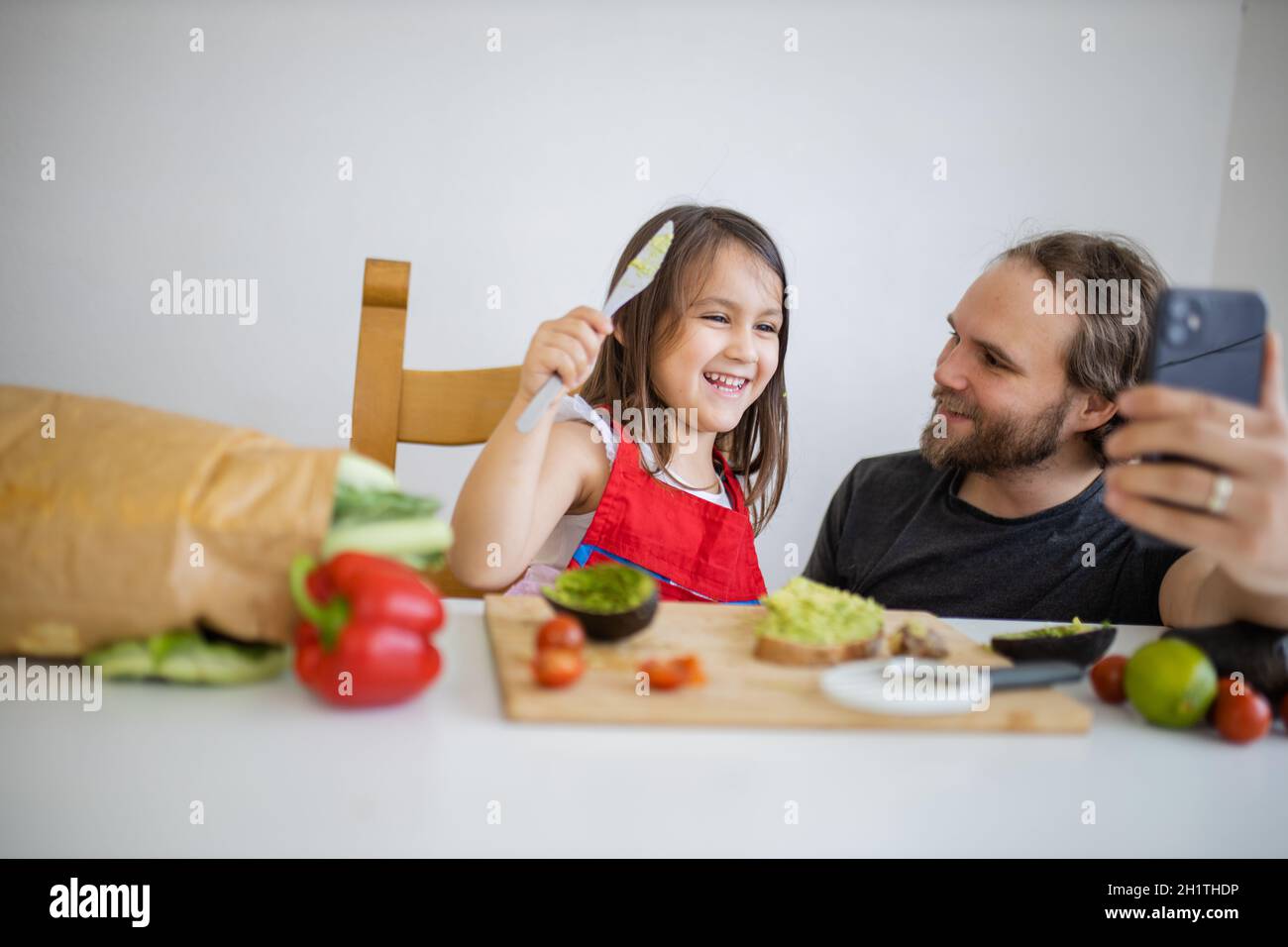 Felice padre e figlia prendere selfie mentre si sparge avocado su toast. Carino bambino che taglia le verdure sul tagliere. Padre-figlia cookin Foto Stock