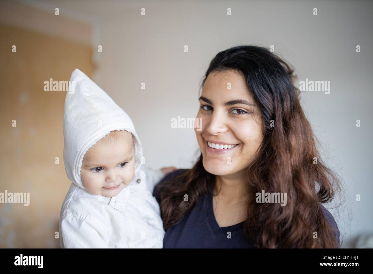 Donna felice sorridendo e tenendo il suo adorabile bambino in abiti bianchi. Ritratto del bambino carino che indossa la felpa con cappuccio bianca e la sua madre gioiosa. Giovane e felice fam Foto Stock