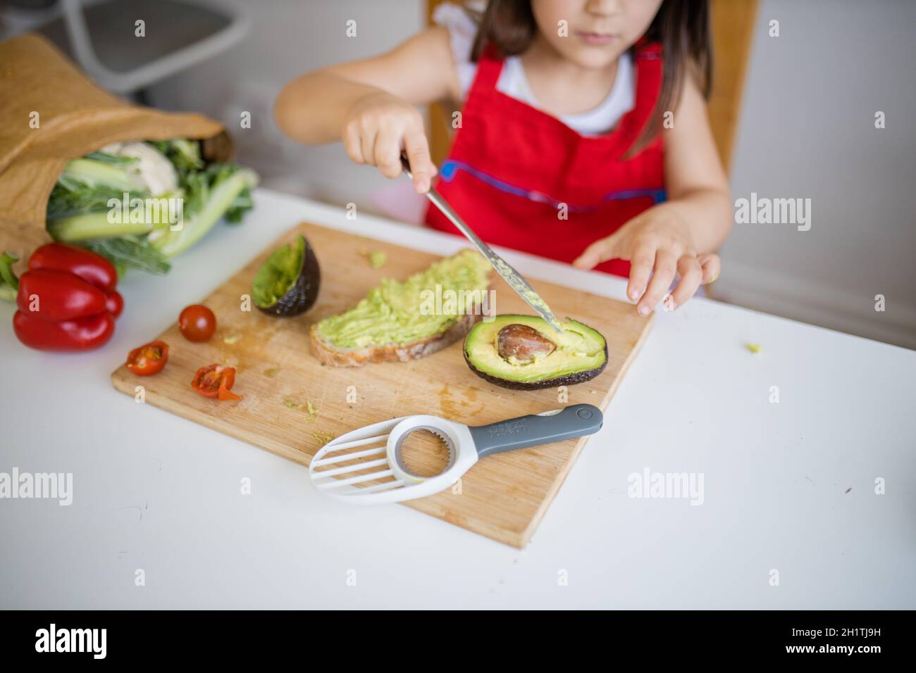 Adorabile bambina al tavolo bianco che diffonde avocado su fetta di pane. Cute e concentrata giovane bambino tagliando le verdure sul tagliere. Bambini p Foto Stock