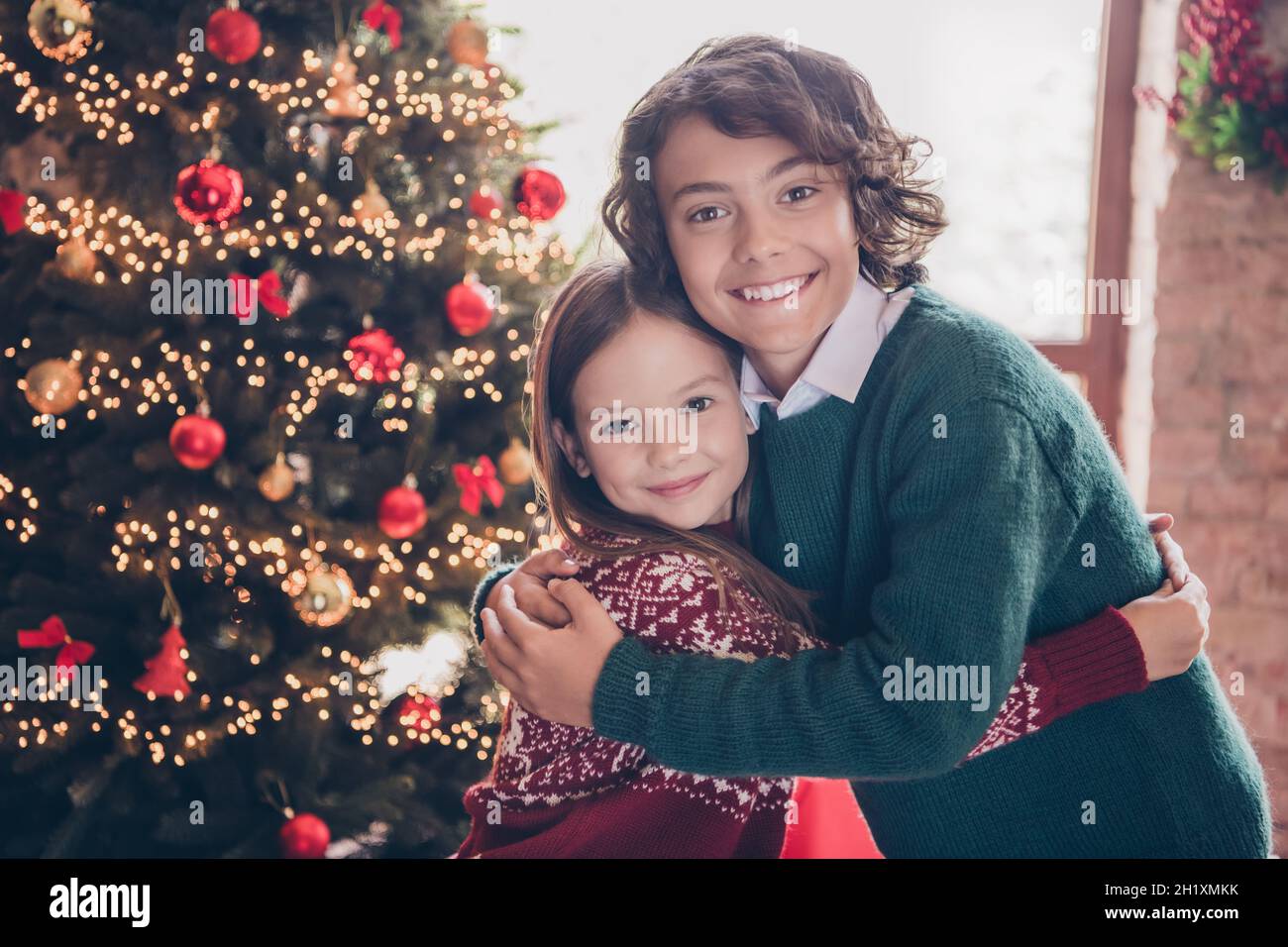 Ritratto piccolo ragazzo e sorella abbracciando vicino ornato albero di Natale in appartamento durante le vacanze invernali Foto Stock