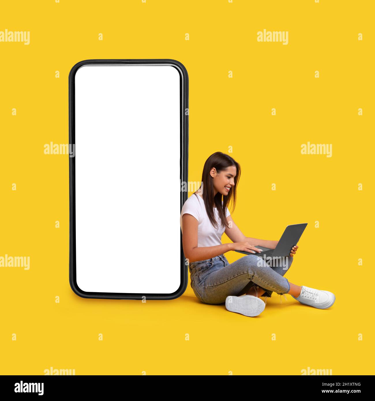 Donna sorridente che usa il laptop, appoggiata sullo schermo bianco vuoto del telefono Foto Stock