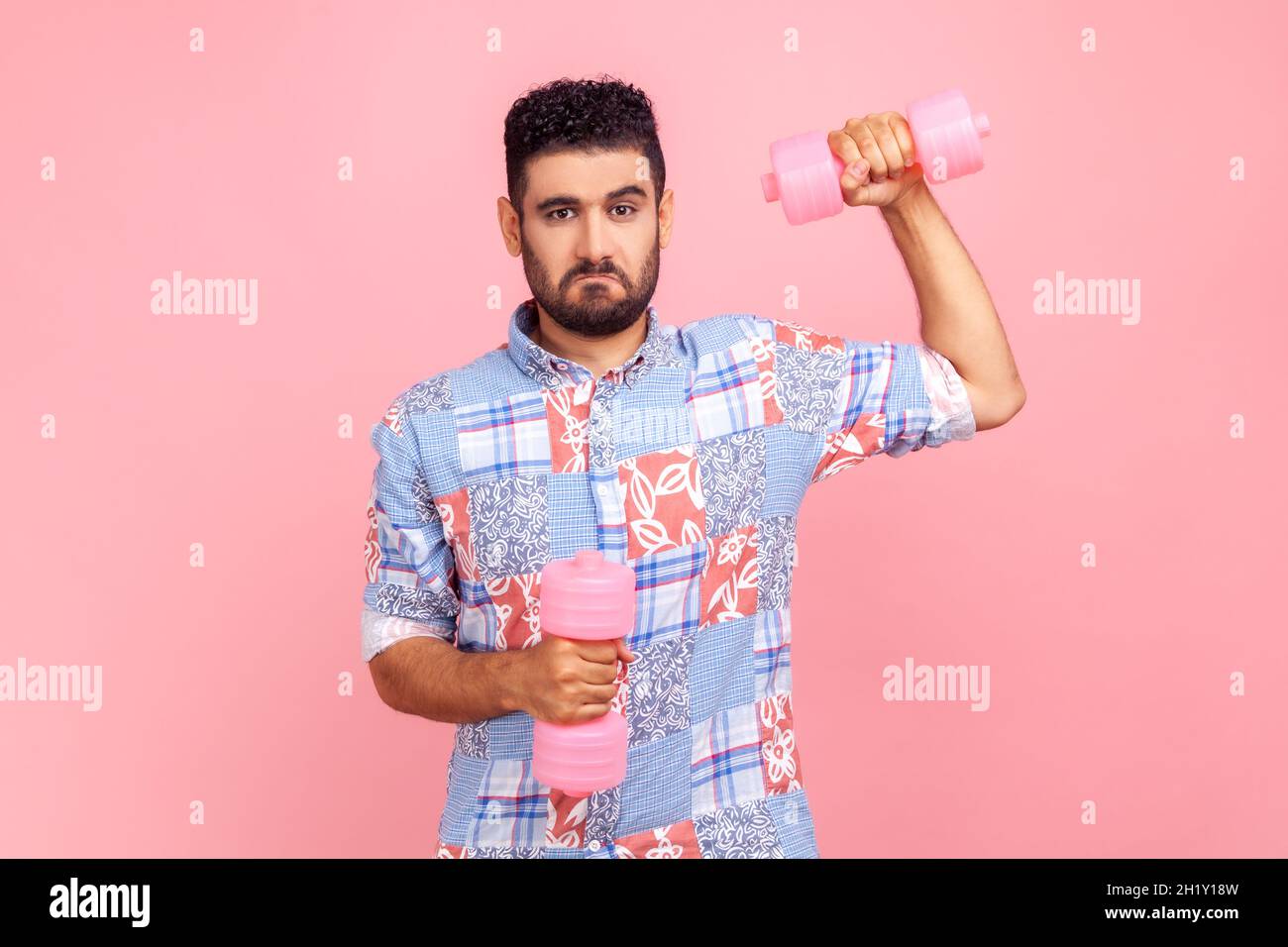 Auto sicuro giovane adulto ported uomo che indossa casual stile camicia blu che tiene manubri, pompando i muscoli, avendo divertente espressione facciale. Studio interno girato isolato su sfondo rosa. Foto Stock