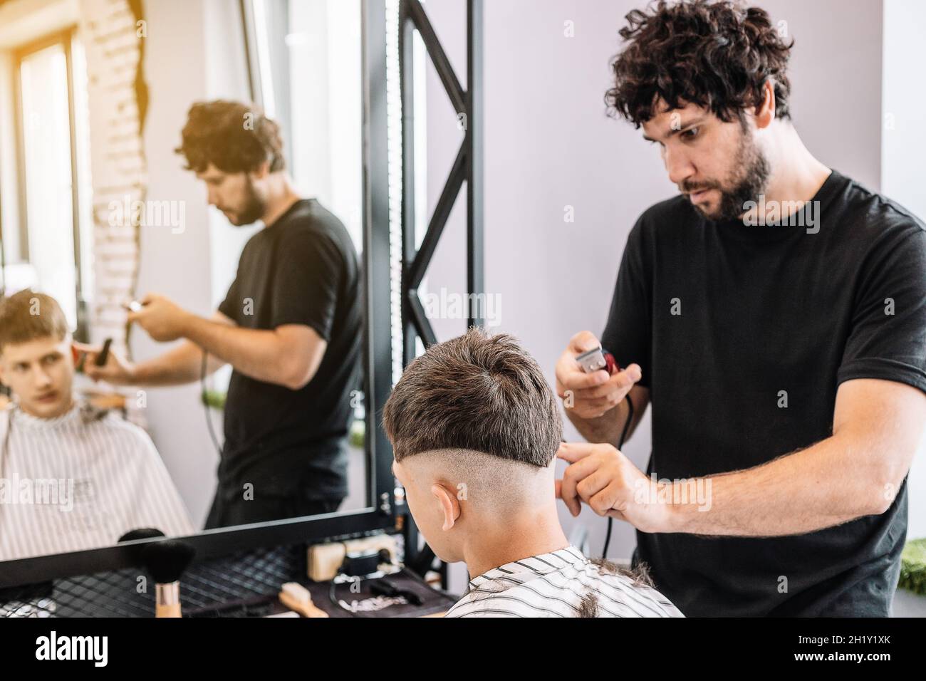 Bello alto barbiere bearded bruna dà un haircut trendy ad un adolescente. Riflessione nello specchio. Taglio professionale e styling per uomo in un barbe Foto Stock