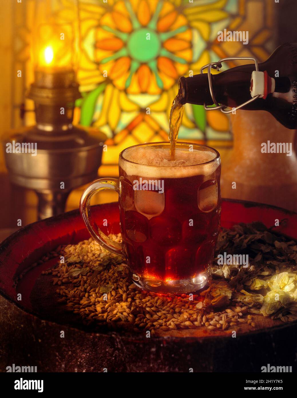 Birra in un bicchiere di pinta con manico versato da una tradizionale bottiglia con tappo ribaltabile sul barile della birra con luppolo e orzo acceso olio lampada e vetro colorato sul retro Foto Stock