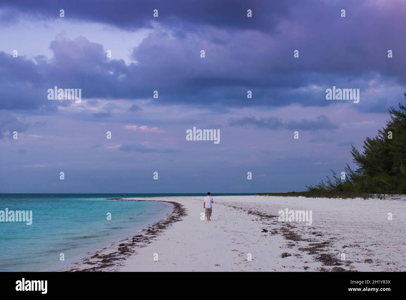 Maschio turista cammina lungo la costa su deserta spiaggia di sabbia bianca sotto il cielo tempestoso al tramonto. Cayo Levisa, Pinar del Rio, Cuba. Foto Stock