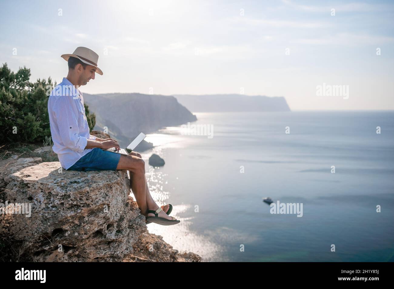 Digital nomad, uomo nel cappello, un uomo d'affari con un laptop siede sulle rocce vicino al mare durante il tramonto, effettua una transazione di affari in linea da a. Foto Stock