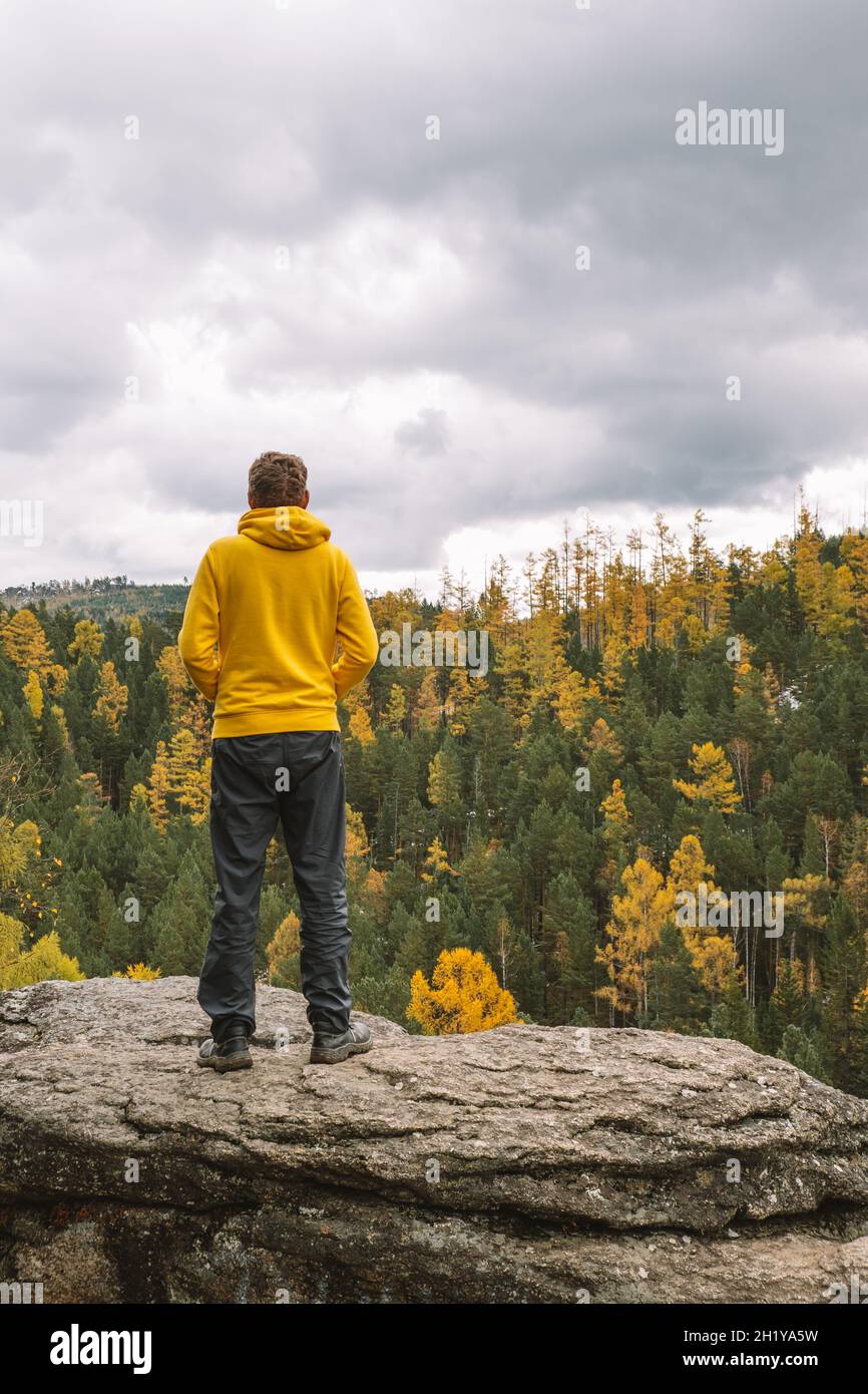 Il viaggiatore maschile in una felpa con cappuccio gialla si erge sulla cima della montagna, ammirando il magnifico paesaggio della foresta gialla d'autunno. Nuvoloso autunno giorno. Foto Stock