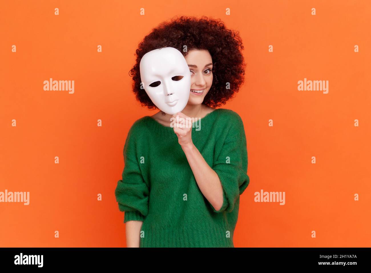 Donna con acconciatura Afro in maglione verde che rimuove la maschera bianca dal volto mostrando la sua espressione sorridente, buon umore, fingendo di essere un'altra persona. Studio interno girato isolato su sfondo arancione. Foto Stock