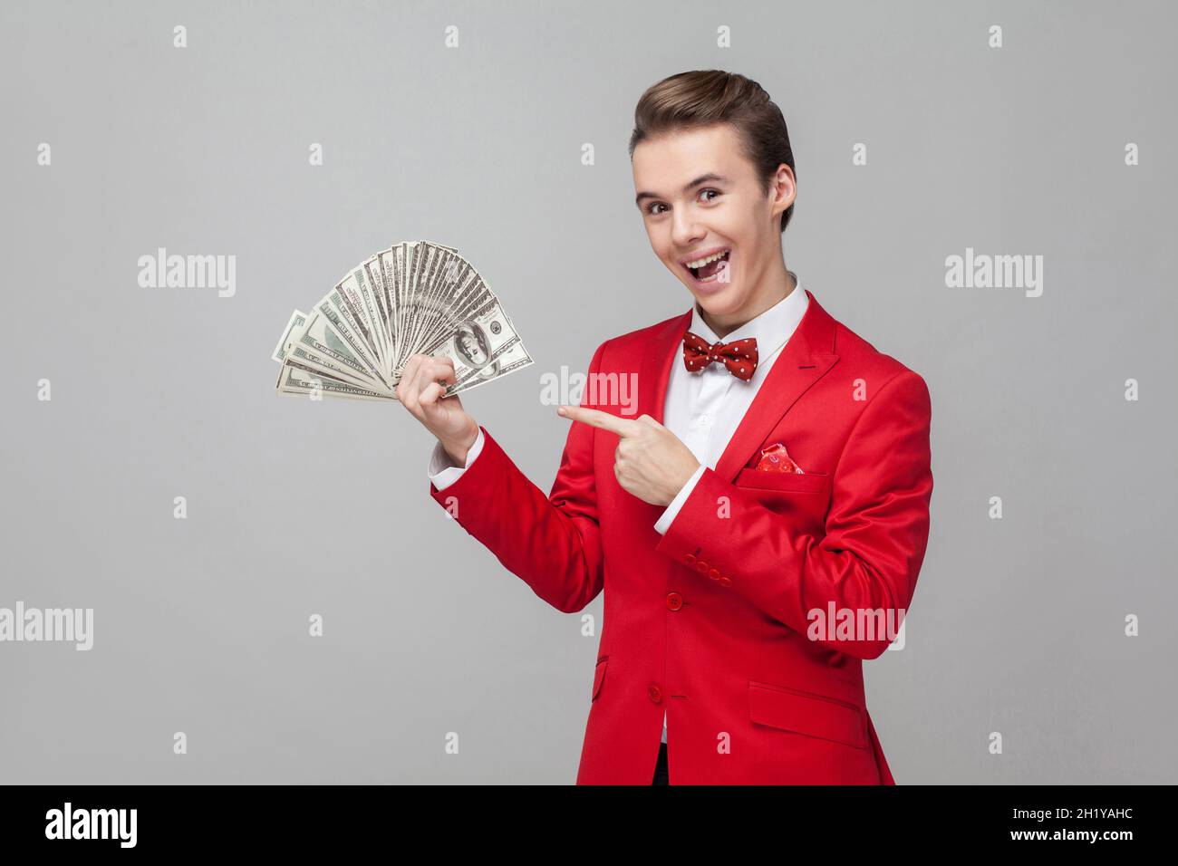 Guarda i miei soldi. Ritratto di uomo estremamente felice con capelli alla moda in giacca rossa e arco cravatta puntando a un mazzo di dollari, esprimendo gioia, eccitazione. studio girato isolato su sfondo grigio Foto Stock