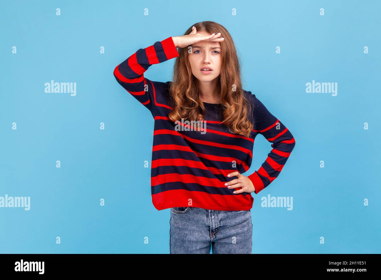 Cercare di vedere qualcosa a distanza. Donna indossando a righe stile casual maglione tiene palmo sulla fronte guarda via cerca qualcuno messo a fuoco lontano . Studio interno girato isolato su sfondo blu. Foto Stock