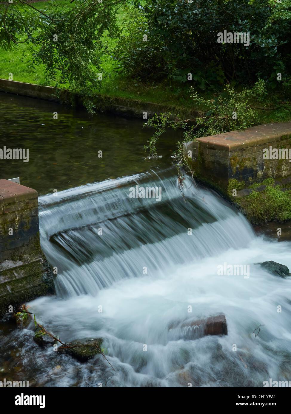 Una piccola cascata di struzzi a più livelli circondata da alberi in un parco suburbano, scattata con un otturatore lento per catturare la susurrazione dell'acqua che cade. Foto Stock