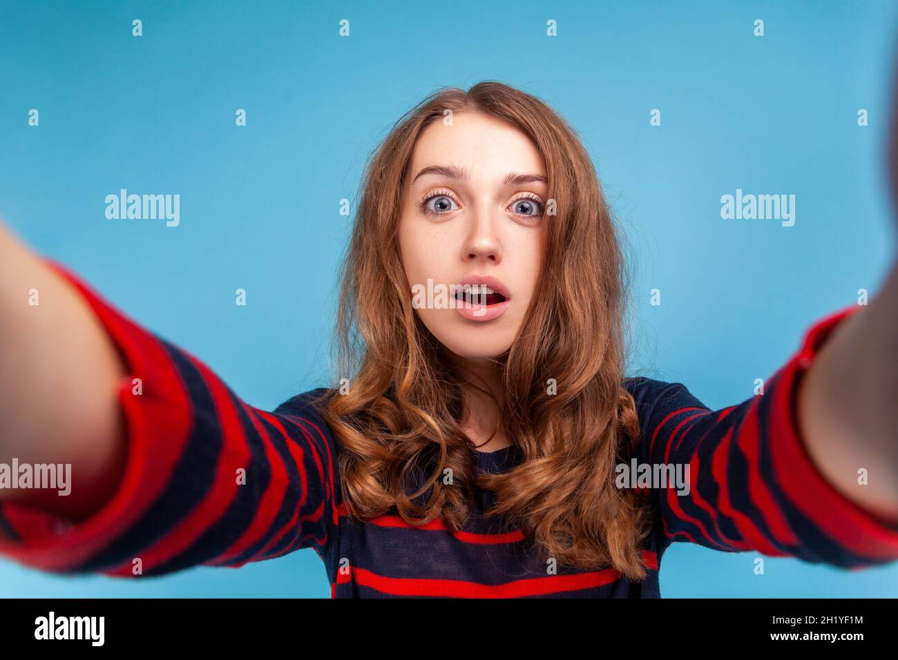 Donna con espressione facciale sorpresa che prende selfie, punto di vista della foto, posa con bocca aperta, indossando un pullover casual a righe. Studio interno girato isolato su sfondo blu. Foto Stock