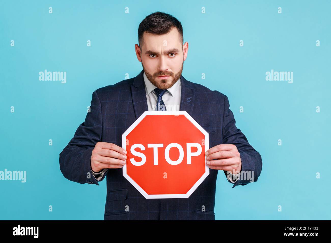 Uomo serio preoccupato che indossa abiti di stile ufficiale tenendo rosso stop segno in mani, attenzione, attenzione concetto attento, avvertenza, guardando la fotocamera. Studio interno girato isolato su sfondo blu. Foto Stock