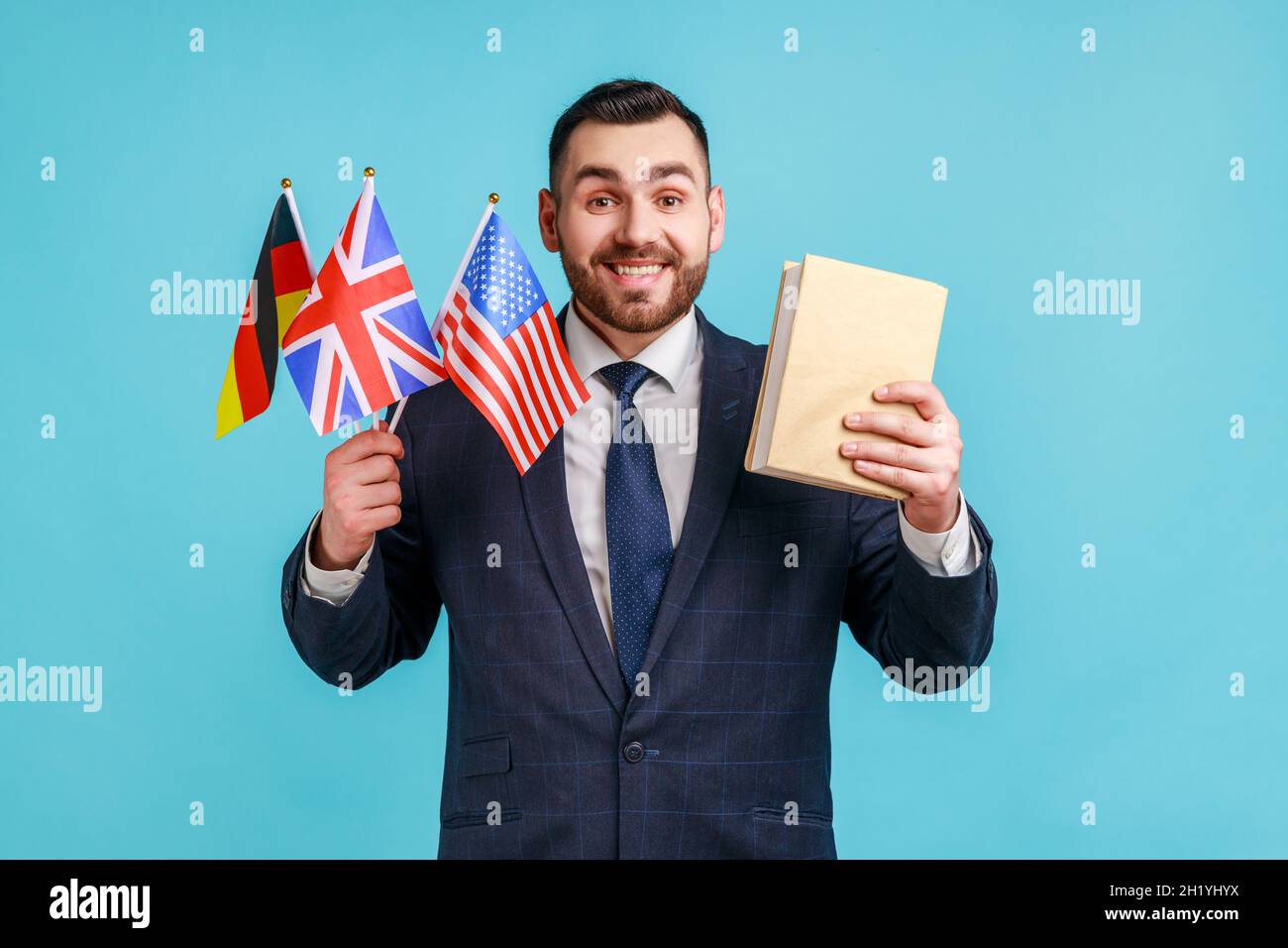 Soddisfatto uomo d'affari o insegnante indossando abiti in stile ufficiale, tenendo bandiere di Germania, Gran Bretagna e Stati Uniti d'America e libro, guardando la macchina fotografica con sorriso. Studio interno girato isolato su sfondo blu. Foto Stock