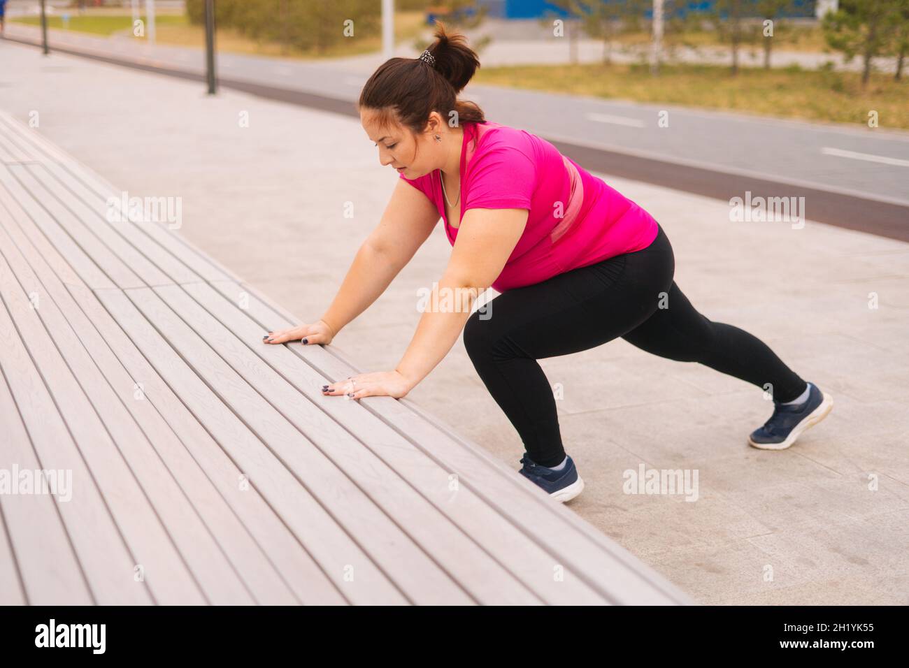 Vista ad alto angolo di motivato grasso giovane donna formazione facendo push up utilizzando panchina strada nel parco della città al mattino d'estate. Foto Stock