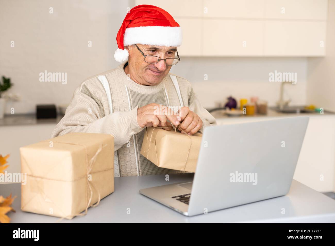 Anziano Gentleman che celebra il Natale in linea al laptop che fa la videochiamata con la famiglia distante seduta in cucina a casa. Capodanno Foto Stock