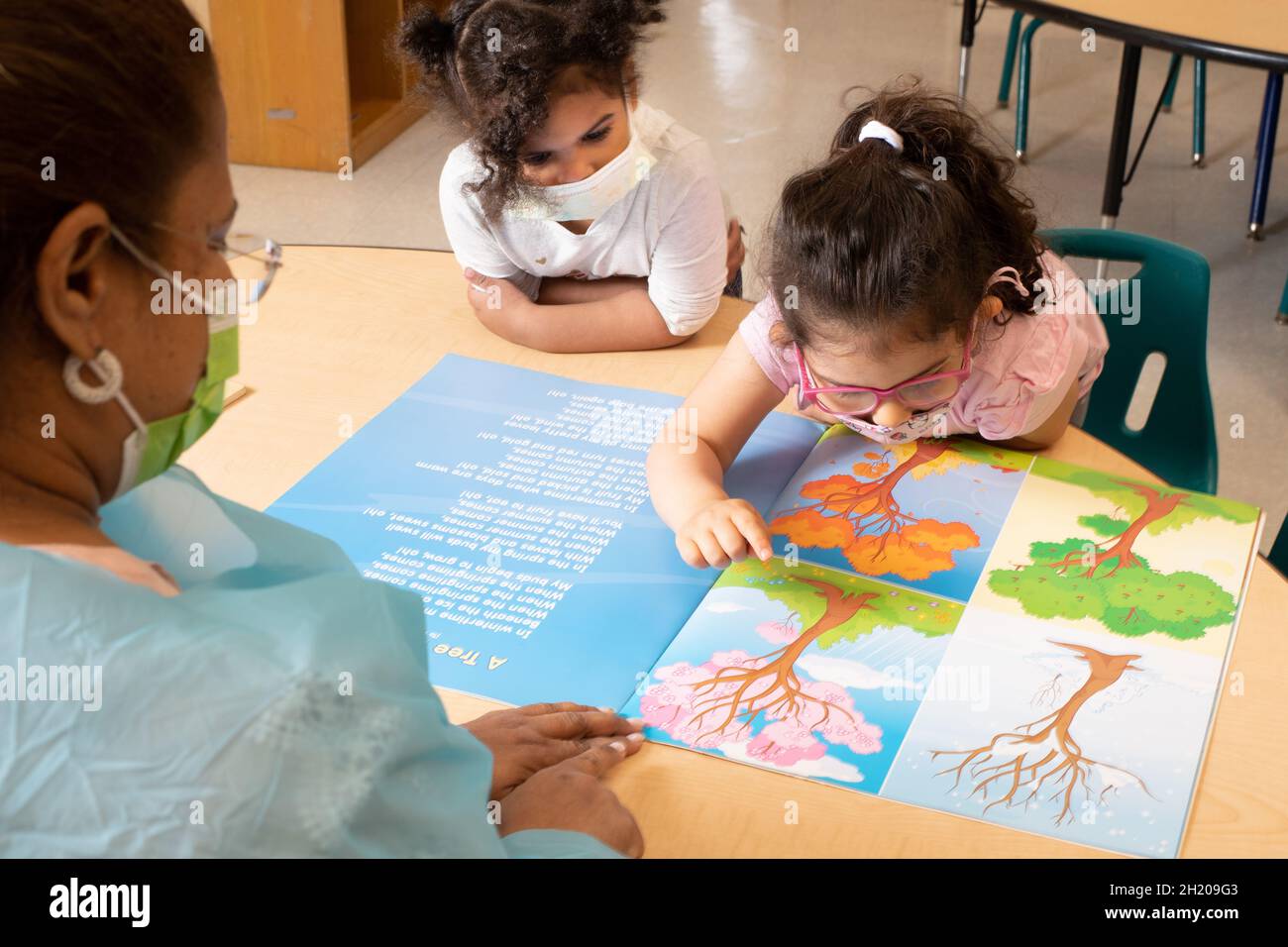 Istruzione Preschool 3-4 anni insegnante guardando un libro di immagini che raffigura le stagioni con due ragazze, un bambino che indica l'immagine dell'albero in sprin Foto Stock