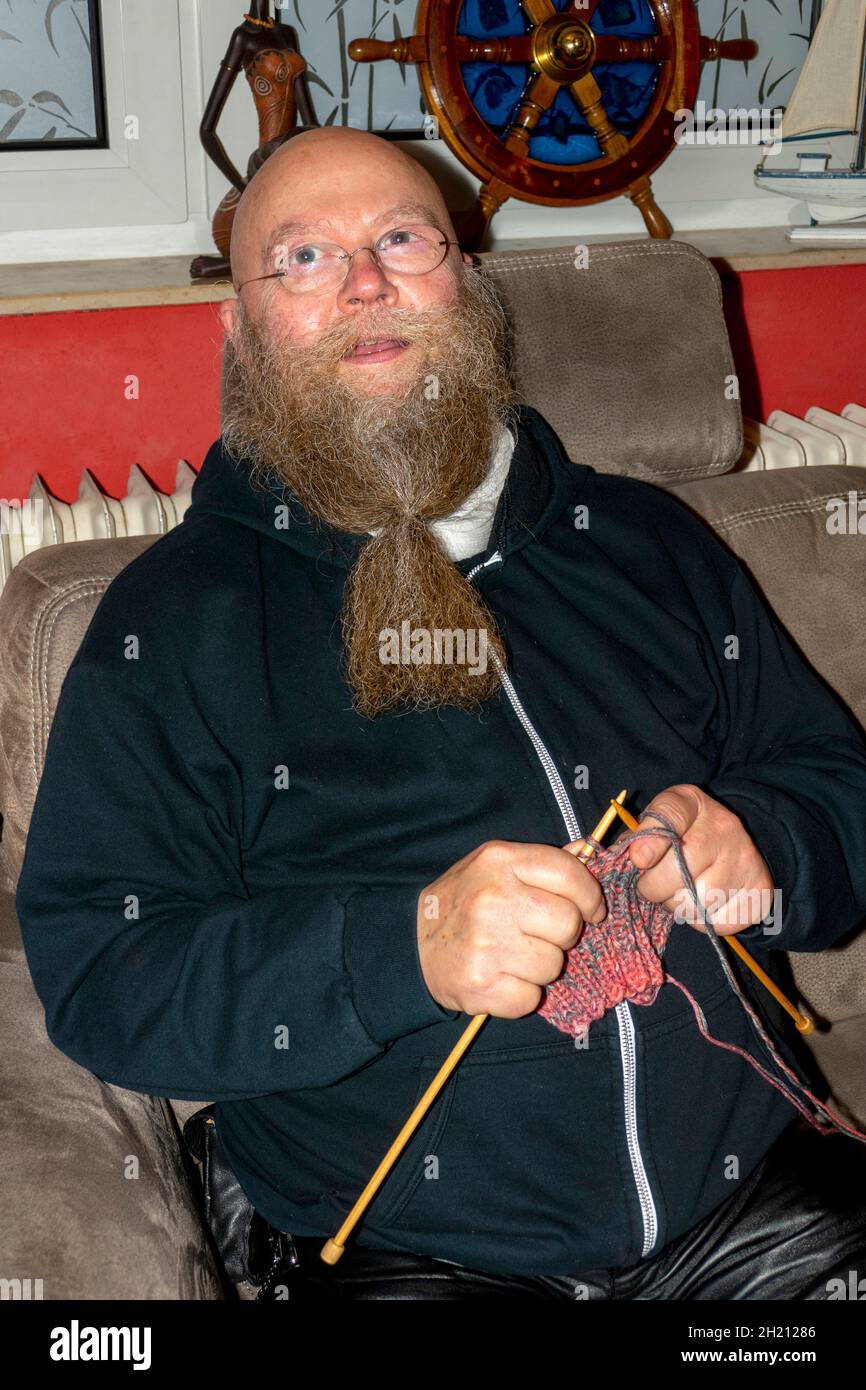 Mann mit Langem Bart und Brille sitzt auf der Couch und strickt einen Schal Foto Stock