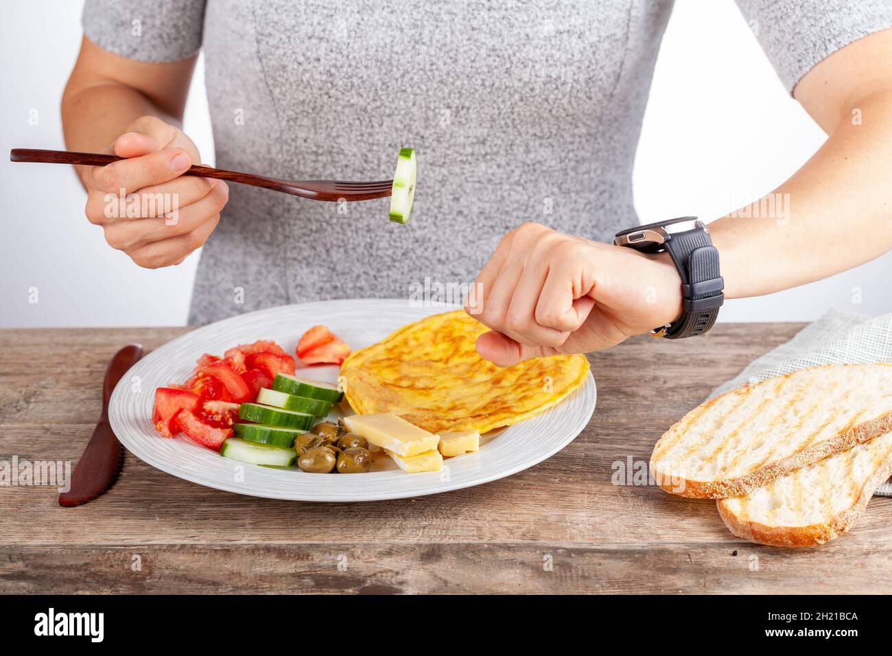 Concetto di digiuno intermittente con una donna che si siede affamata di fronte al cibo e guarda il suo orologio per assicurarsi che si rompa veloce sul momento giusto. R Foto Stock