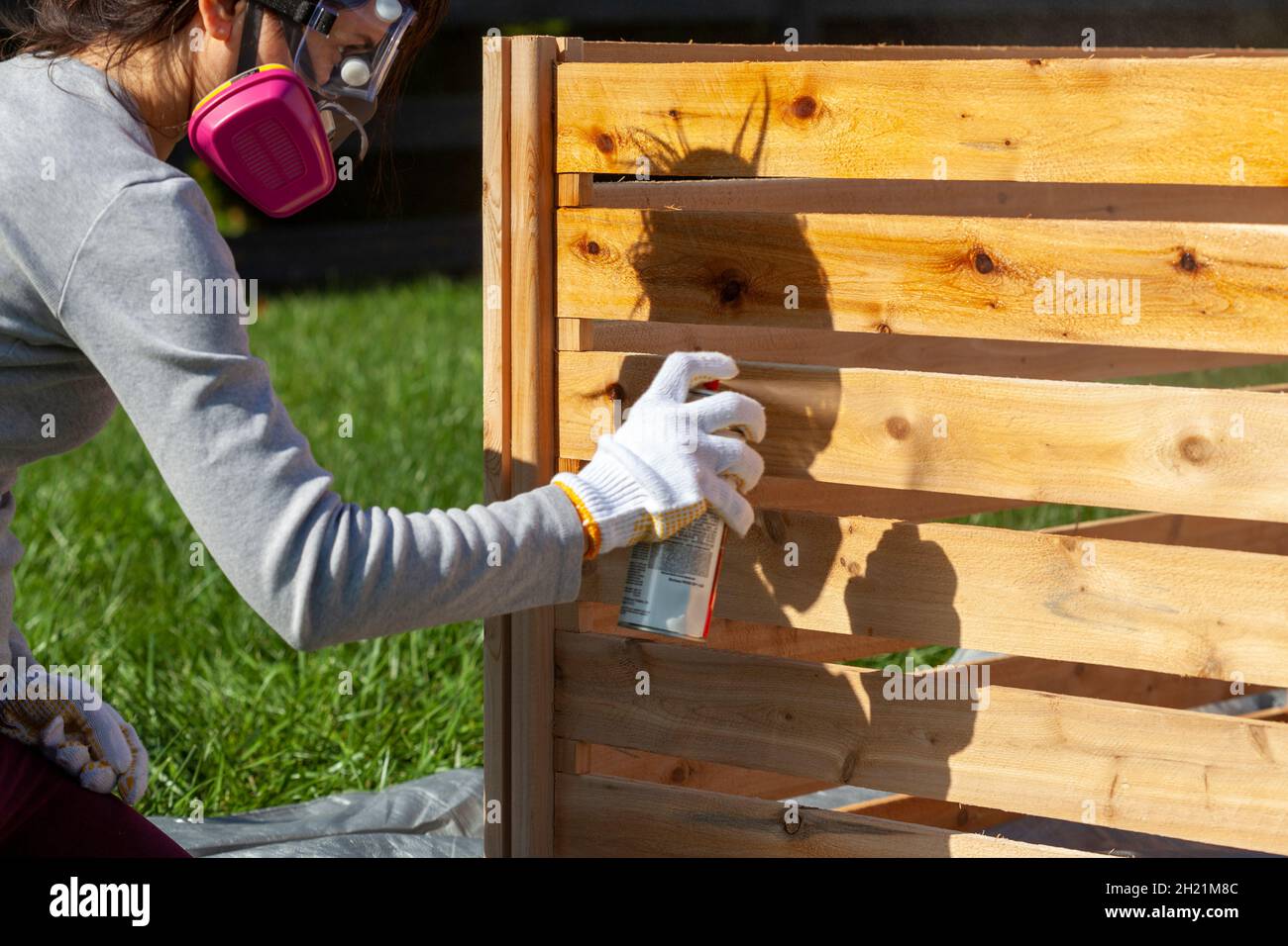 Una giovane donna caucasica che indossa maschera di protezione occhiali e guanti è spray macchia una recinzione di legno all'aperto. Immagine concettuale per il progetto diy e la riparazione Foto Stock