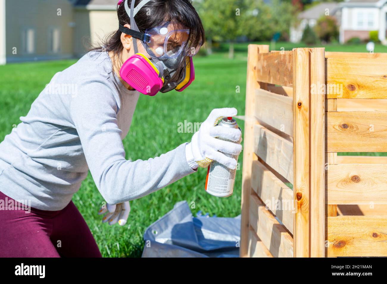 Una giovane donna caucasica che indossa maschera di protezione occhiali e guanti è spray macchia una recinzione di legno all'aperto. Immagine concettuale per il progetto diy e la riparazione Foto Stock