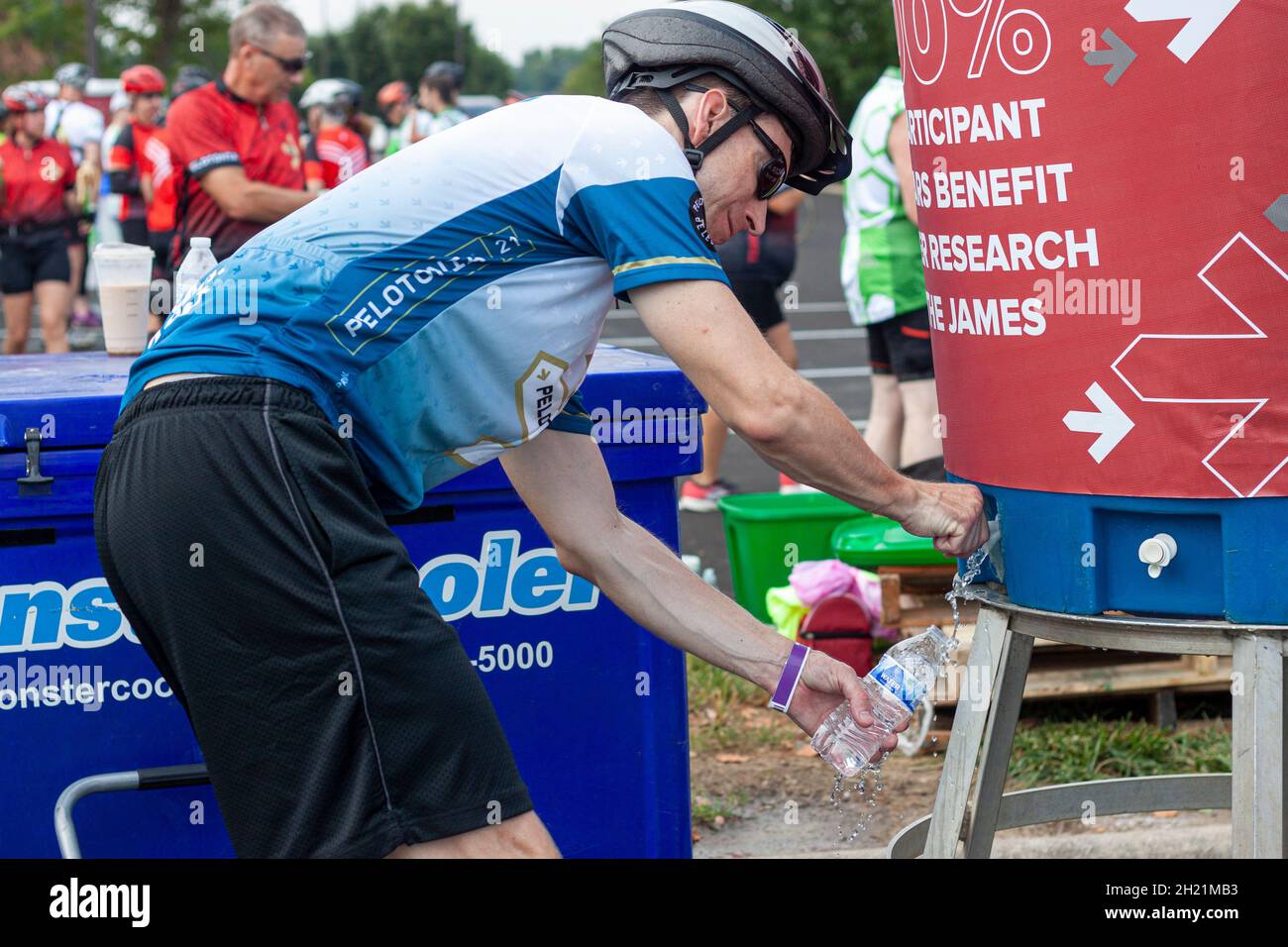 Columbus, OH USA 08-07-2021: Un uomo caucasico che indossa il casco da ciclismo sta riempiendo la sua bottiglia d'acqua di plastica da un grande serbatoio di stoccaggio dell'acqua durante un sp Foto Stock