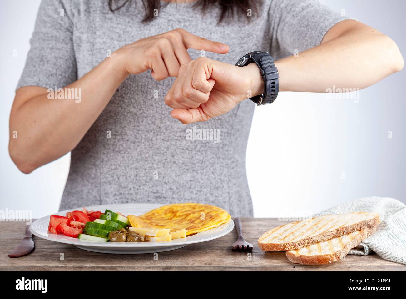 Concetto di digiuno intermittente con una donna che si siede affamata di fronte al cibo e guarda il suo orologio per assicurarsi che si rompa veloce sul momento giusto. R Foto Stock