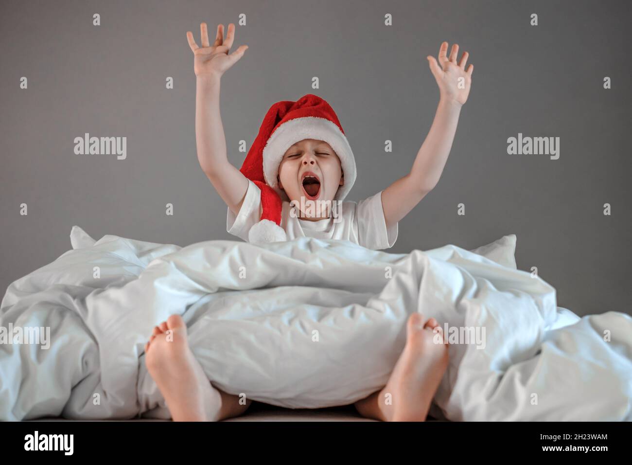 Il ragazzo assonnato è seduto sul letto sorseggiando un cappello di Santa. Capodanno e festività. Felicità mattina di Natale Foto Stock