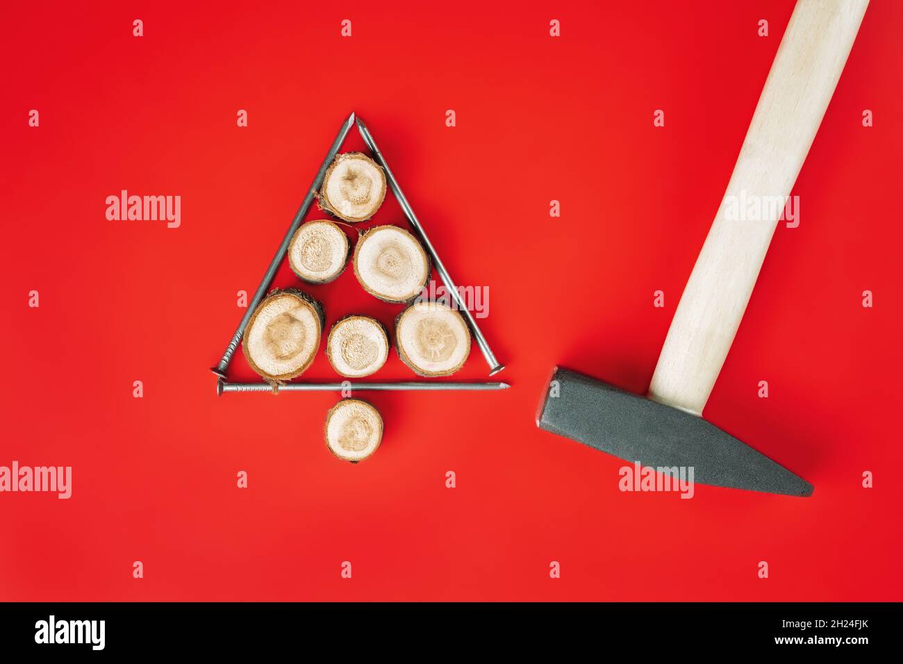 Chiodi lunghi e tagli di legno o lastre di ramo, disposti a forma di albero di Natale con martello su sfondo rosso. Disposizione piatta Foto Stock