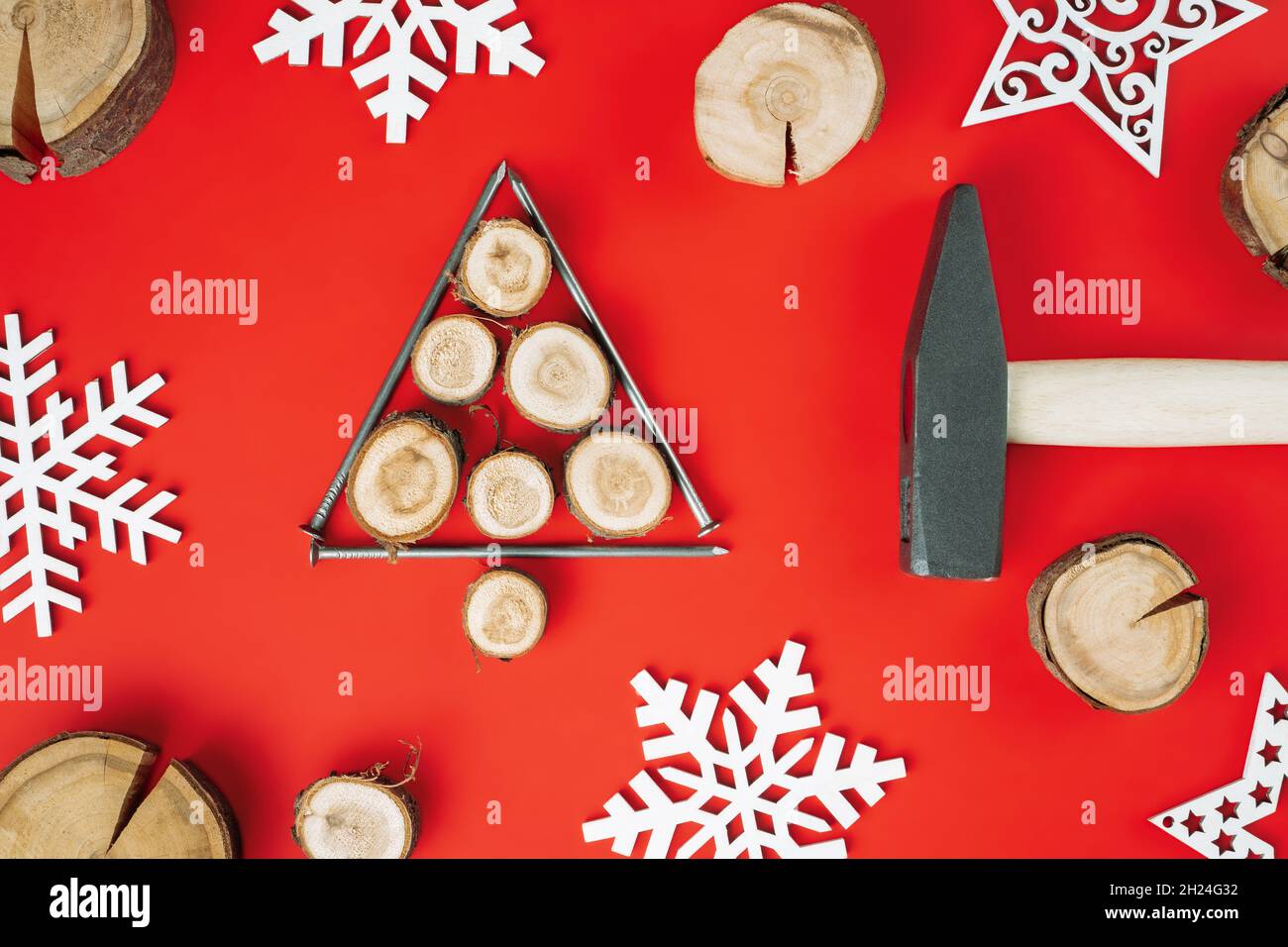 Chiodi lunghi e tagli di legno o lastre di ramo, disposti a forma di albero di Natale con martello e fiocchi di neve in legno su sfondo rosso. Disposizione piatta Foto Stock