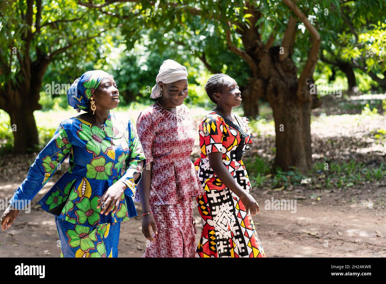 Tre belle giovani donne nere con colorati abiti festosi che camminano fianco a fianco su un sentiero forestale, cantando una canzone tradizionale africana Foto Stock