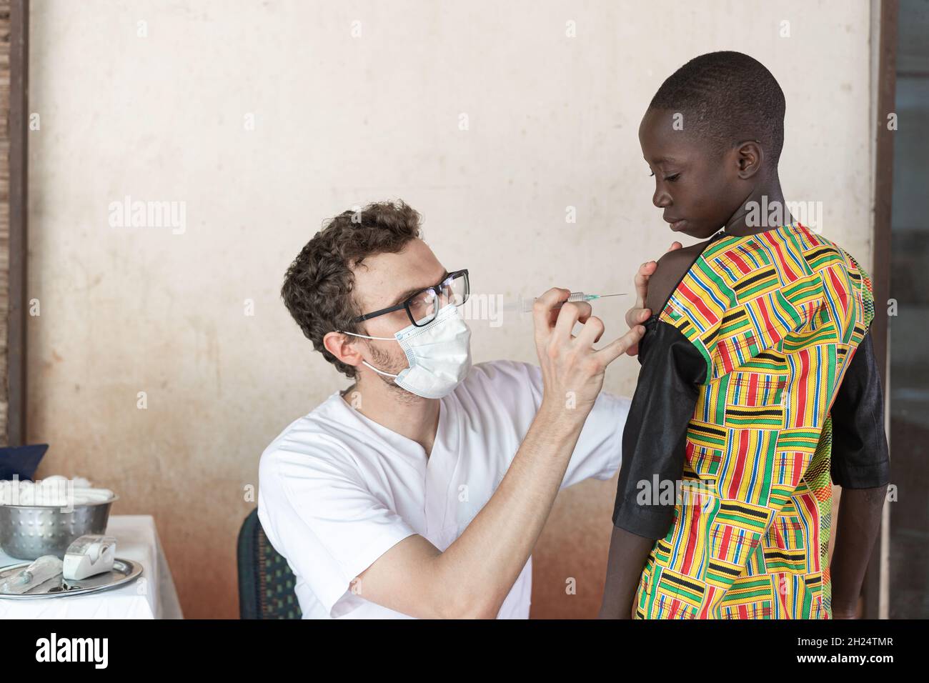 Medico con maschera protettiva per il viso preparazione del sito di iniezione sul braccio di un coraggioso scolaro africano nero in oder per somministrare un f Foto Stock