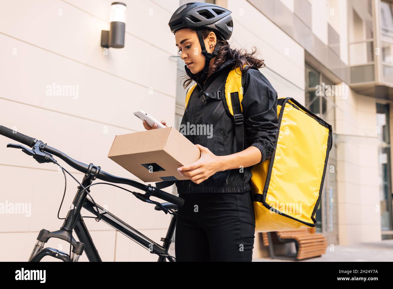 Vista laterale della donna che indossa casco da bicicletta e zaino che guarda sul telefono cellulare mentre tiene un pacco Foto Stock