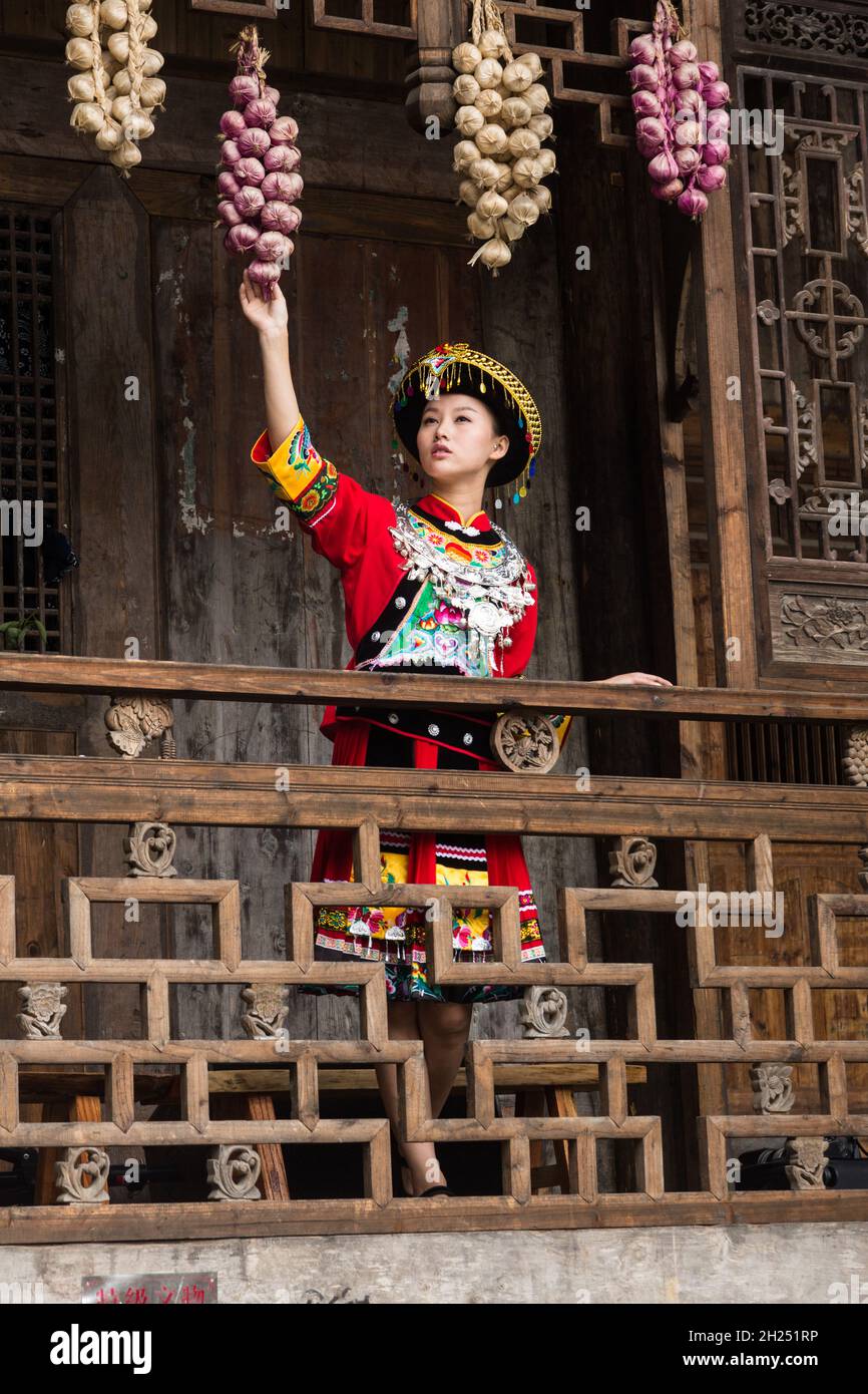 Una giovane donna attraente in tradizionale abito Tujia etnica pone per un ritratto a Furong, Cina. Fasci di cipolle colorate appendono dalle gronde come d Foto Stock