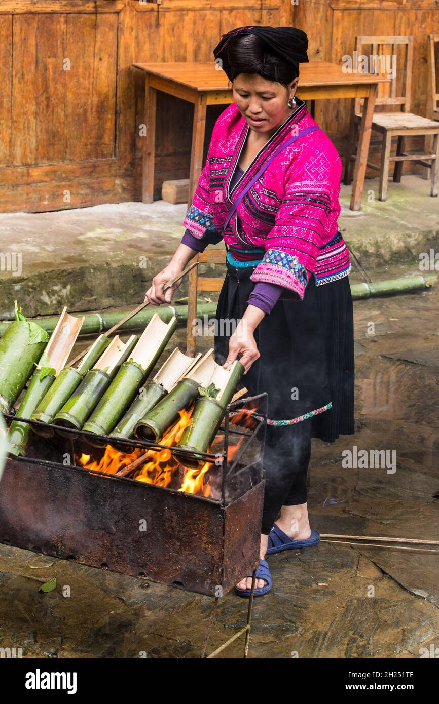 Una donna di Yao rosso che cucinava riso in tubi di bambù su un fuoco aperto a Jinking, Longshen, Cina. Foto Stock