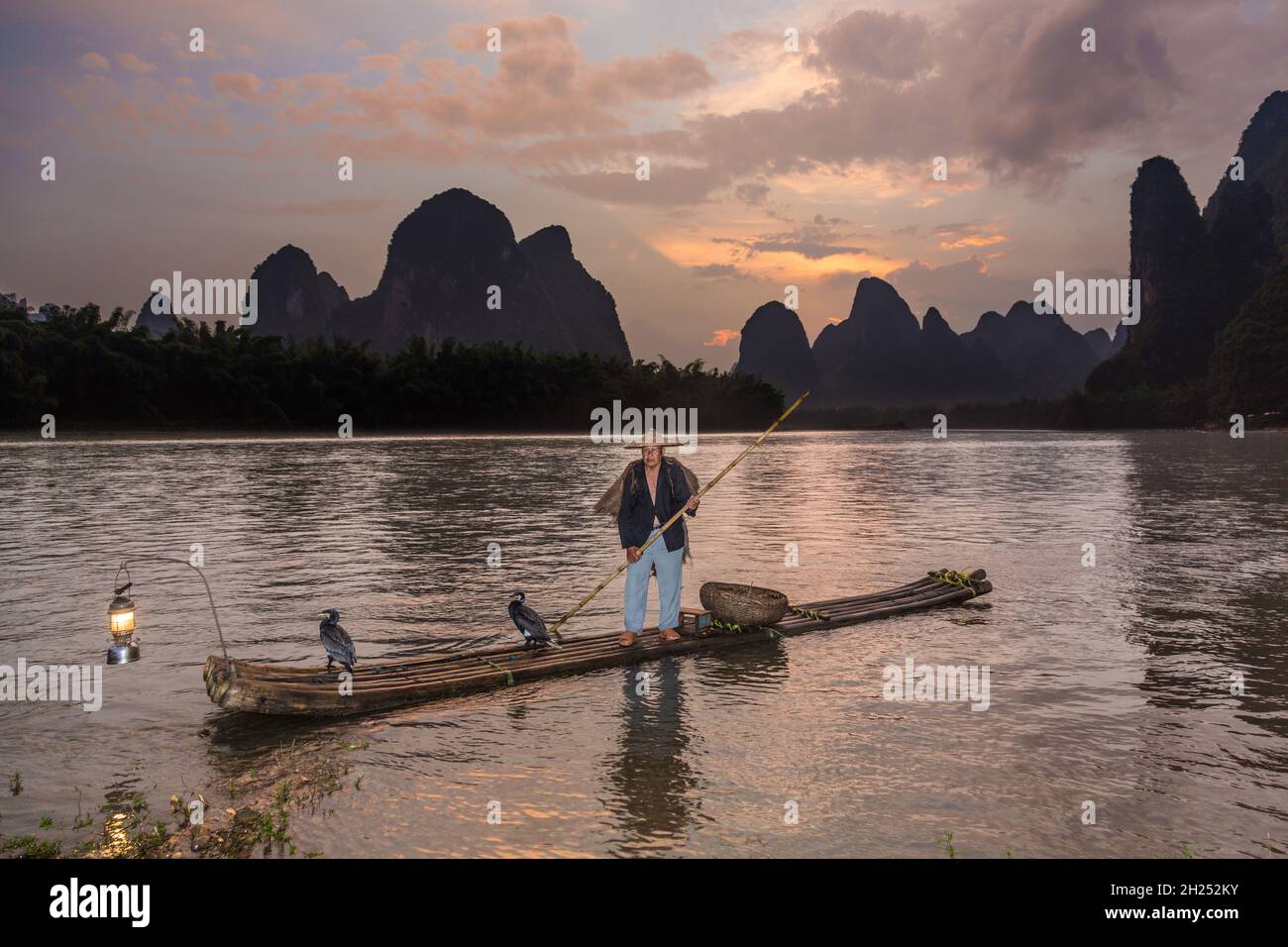 Pescatore cormorano in cappello conico e mantello su zattera di bambù con cormorani e lanterna. Li River, Cina. Foto Stock