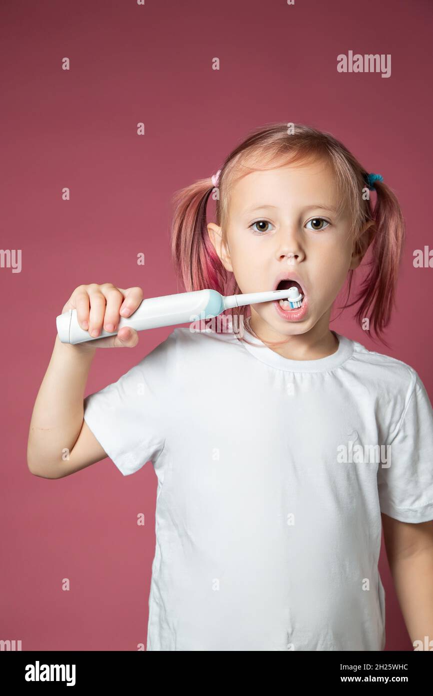 Sorridente bambina caucasica che pulisce i denti con lo spazzolino elettrico sonico su sfondo rosa. Foto Stock