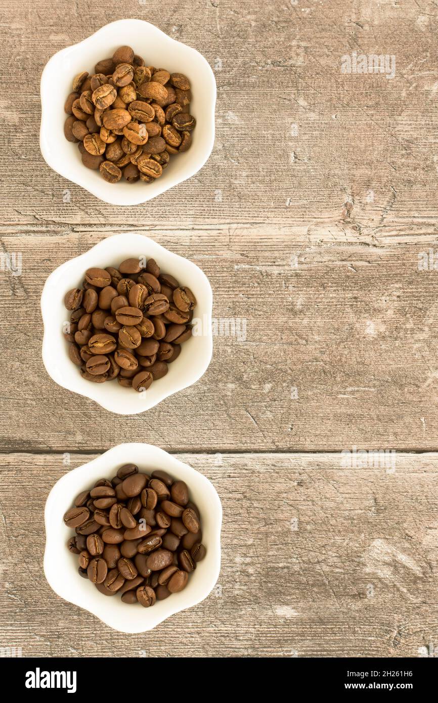 Tostatura leggera, tostatura media e tostatura scura, fagioli arabica in ciotole bianche su legno intemperiato marrone con spazio copia. Vista dall'alto. Foto Stock