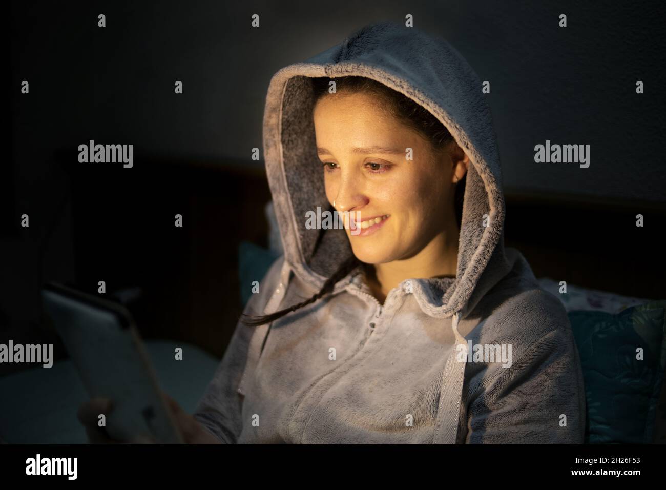 Primo piano di una giovane donna in una felpa con cappuccio seduta nell'oscurità e guardando la tavoletta. Foto Stock
