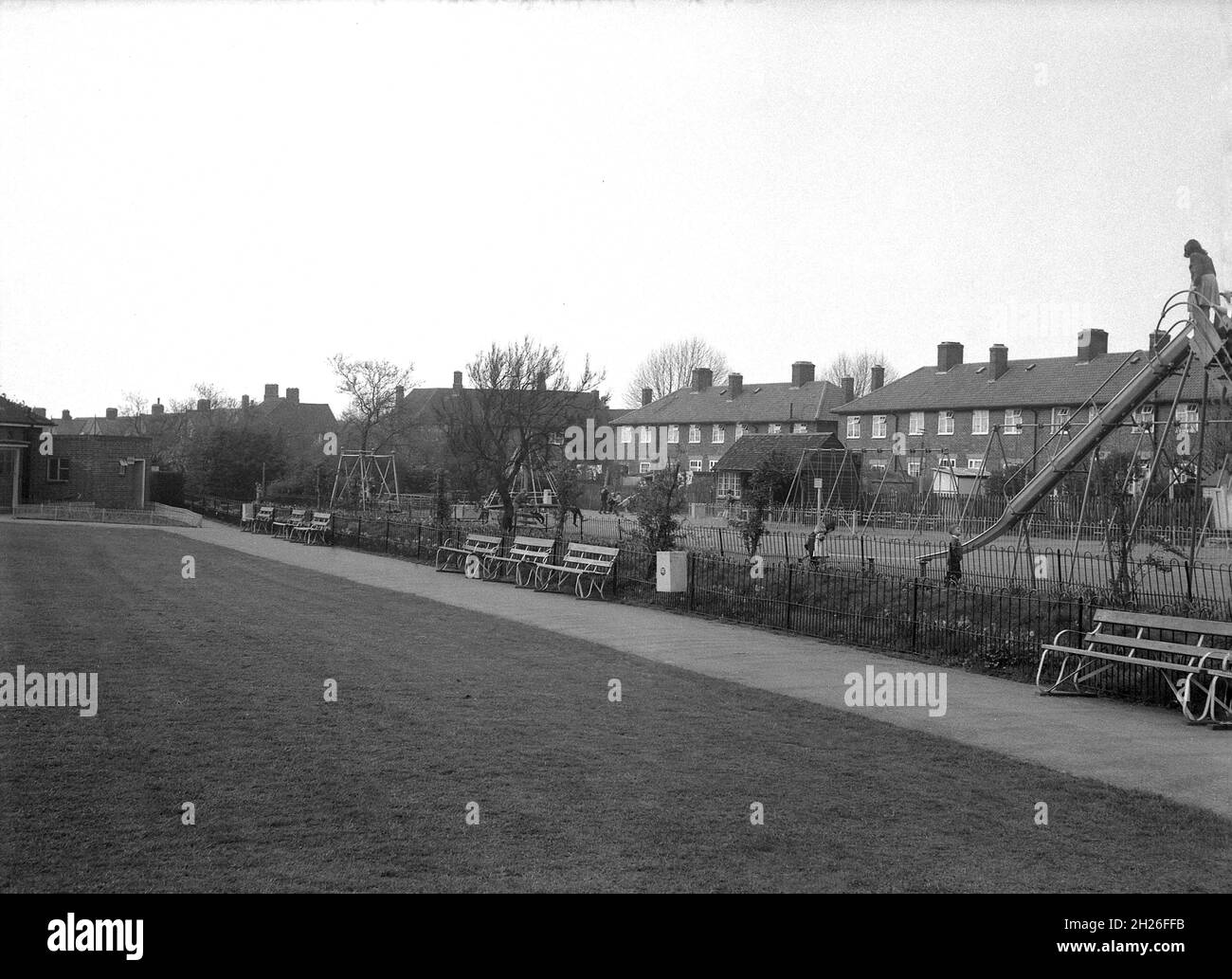 Anni '50, storico, vista di un parco pubblico suburbano con campo sportivo situato da una fila di case locali, Inghilterra, Regno Unito. I bambini possono essere visti giocare accanto al parco in un'area giochi con attrezzature tradizionali, tra cui uno scivolo di metallo, una capanna delle streghe e una rotatoria, forse Abbey Wood o Eltham nel sud di Londra. Foto Stock