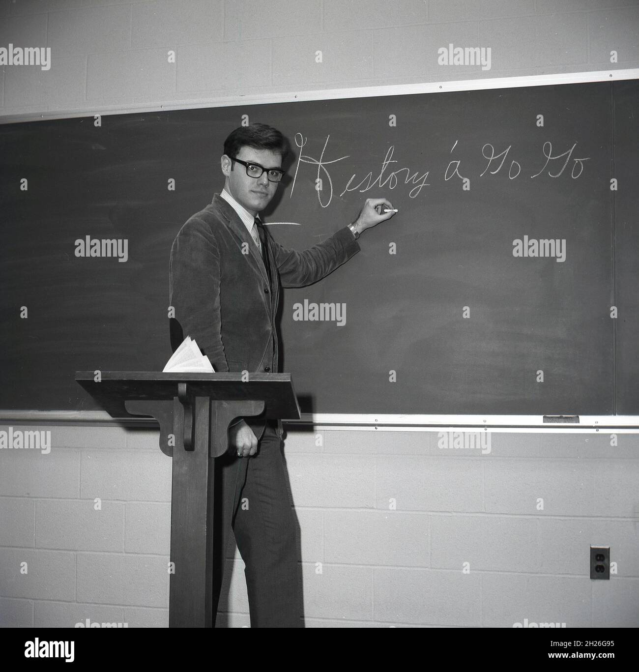 Anni '60, storico, un giovane insegnante di sesso maschile in un abito corduroy e cravatta in piedi dietro un leggio in una lavagna con gesso in mano, Monroe College, Middletown, Virginia, USA Foto Stock
