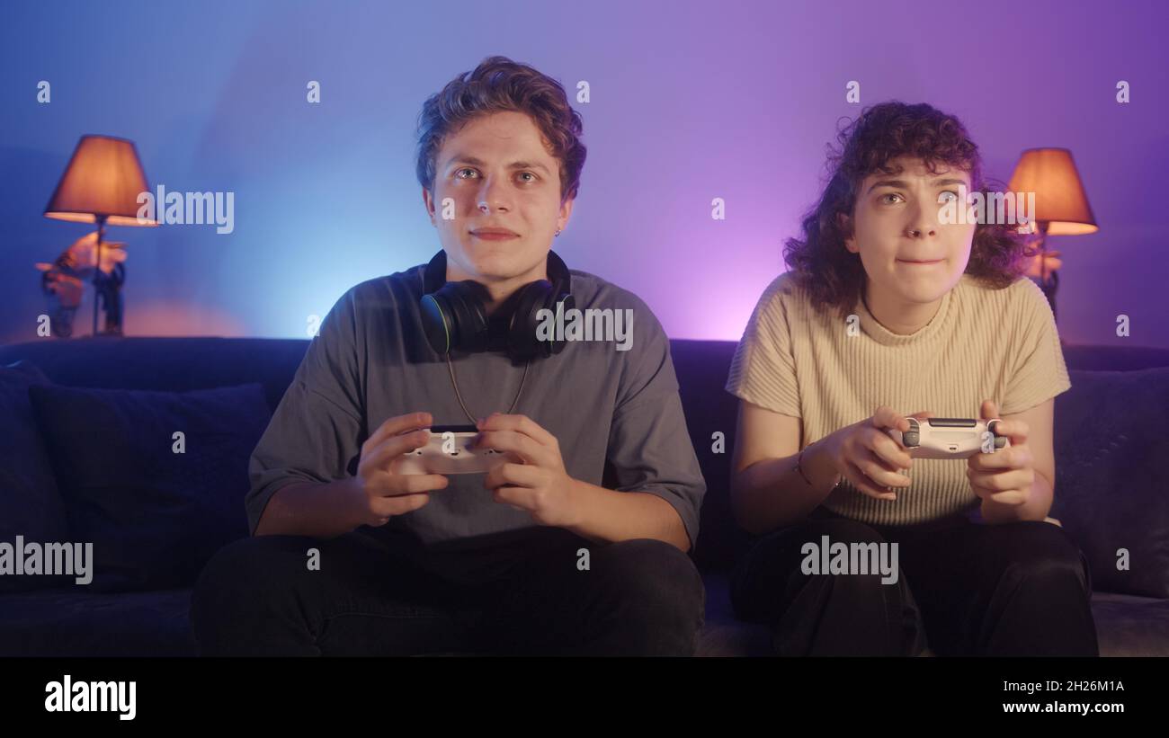 Cute curly-hair ragazza e bel ragazzo biondo sono seduti sul divano a casa e giocare un video gioco su console Foto Stock